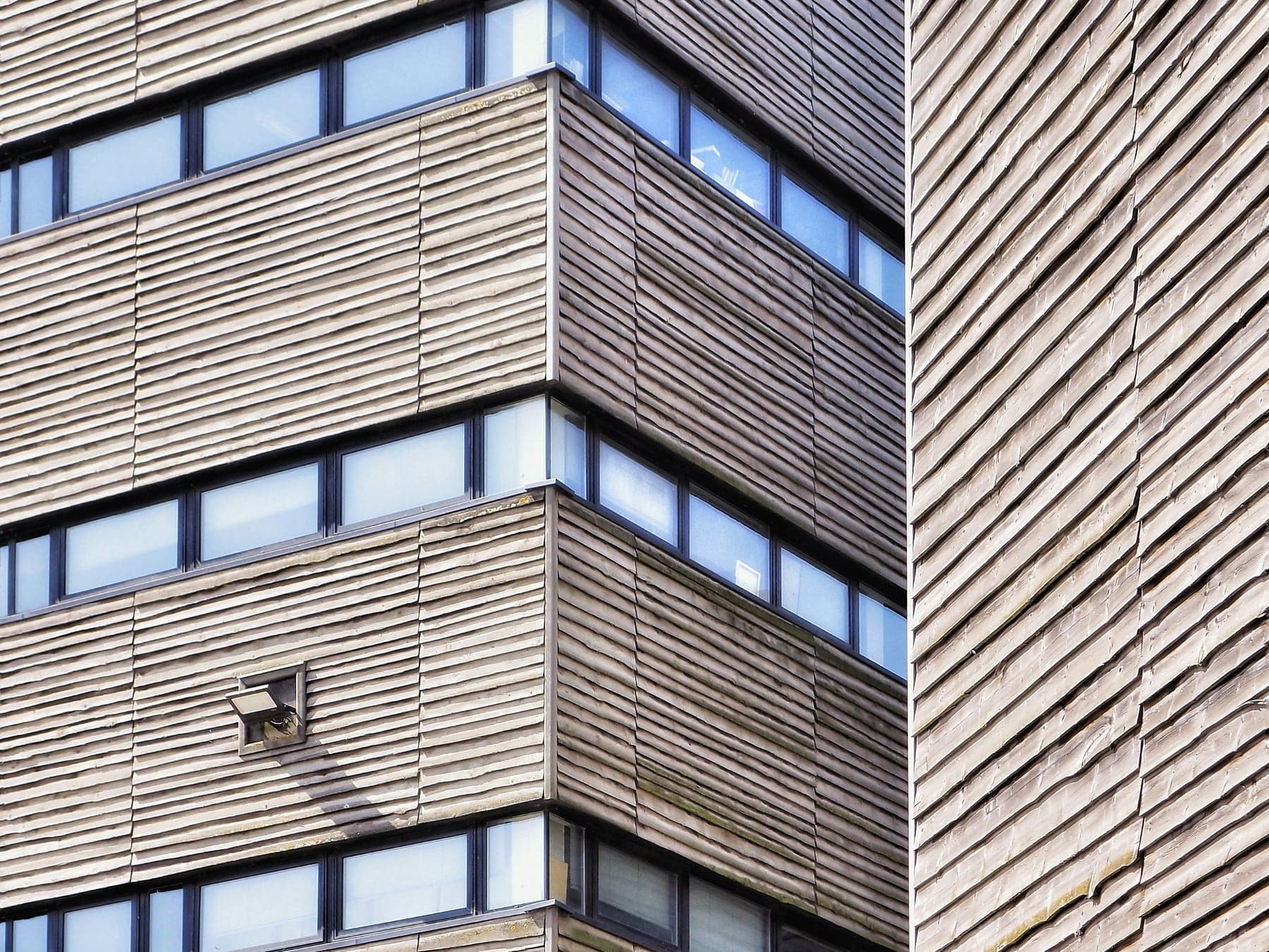 Ecke eines modernen Verwaltungsgebäudes aus Sichtbeton mit vorgehängten waagerechten Holzlamellen und Fensterbändern.